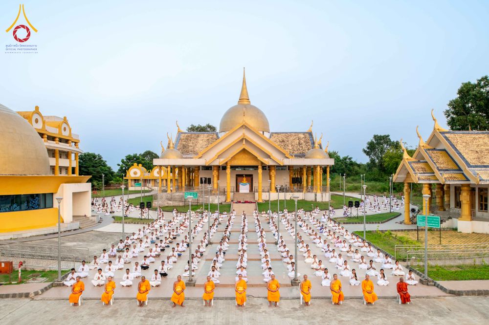 พระภิกษุและสาธุชนจำนวนมากนั่งสมาธิเรียงแถวอย่างเป็นระเบียบหน้าอาคารสถาปัตยกรรมสีทอง