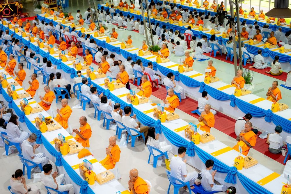 คณะสงฆ์และสาธุชนจำนวนมากนั่งเรียงรายตามโต๊ะยาวภายในอาคารเพื่อร่วมงานบุญ