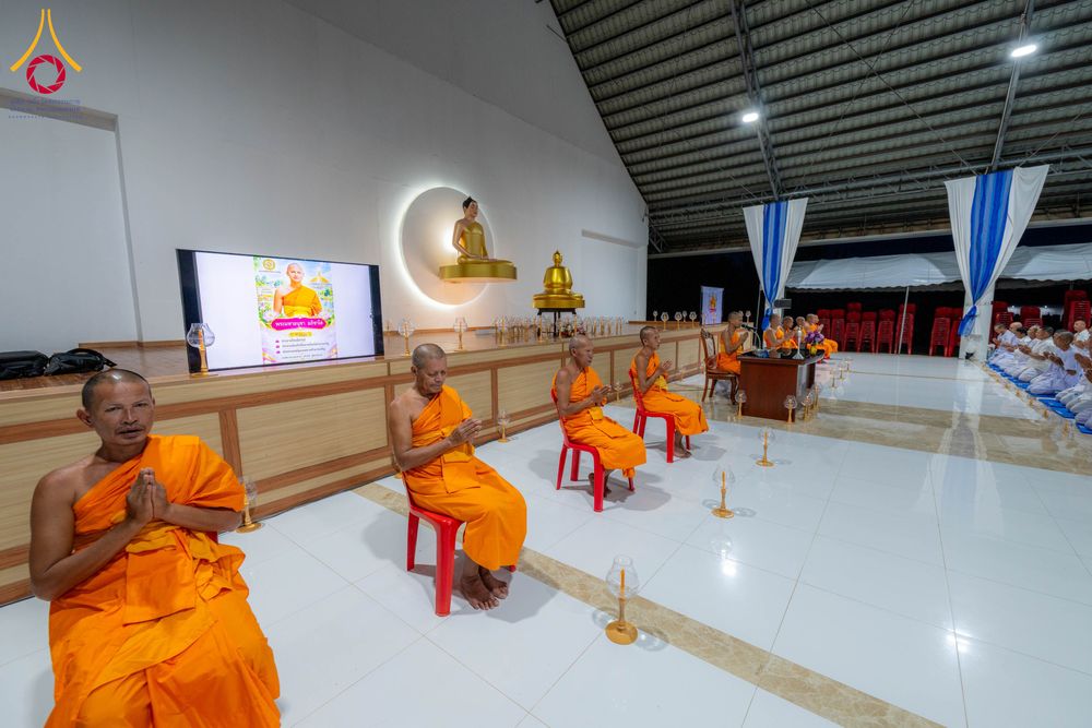 พระภิกษุสงฆ์นั่งพนมมือปฏิบัติธรรมภายในอาคารที่มีพระพุทธรูปและจอภาพแสดงข้อมูลโครงการ