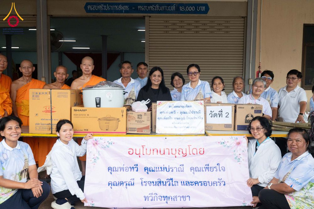 กลุ่มพระภิกษุและคณะผู้มีจิตศรัทธายืนเรียงแถวหน้าโต๊ะที่มีกล่องเครื่องอุปโภคบริโภคและป้ายข้อความอนุโมทนาบุญ