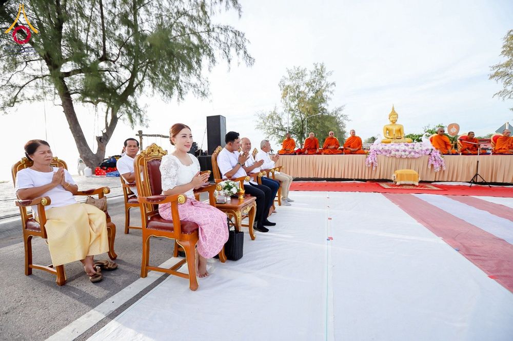 กลุ่มคนนั่งพนมมือร่วมพิธีทางศาสนาบริเวณริมชายหาดโดยมีพระสงฆ์นั่งอยู่บนเวทีด้านหน้า