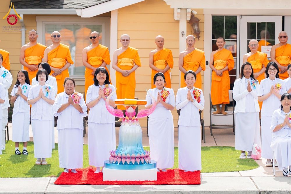 พระภิกษุสงฆ์และกลุ่มสาธุชนชุดขาวกำลังประกอบพิธีทางศาสนาพุทธ โดยมีพานพุ่มผ้าไตรตั้งอยู่ด้านหน้า