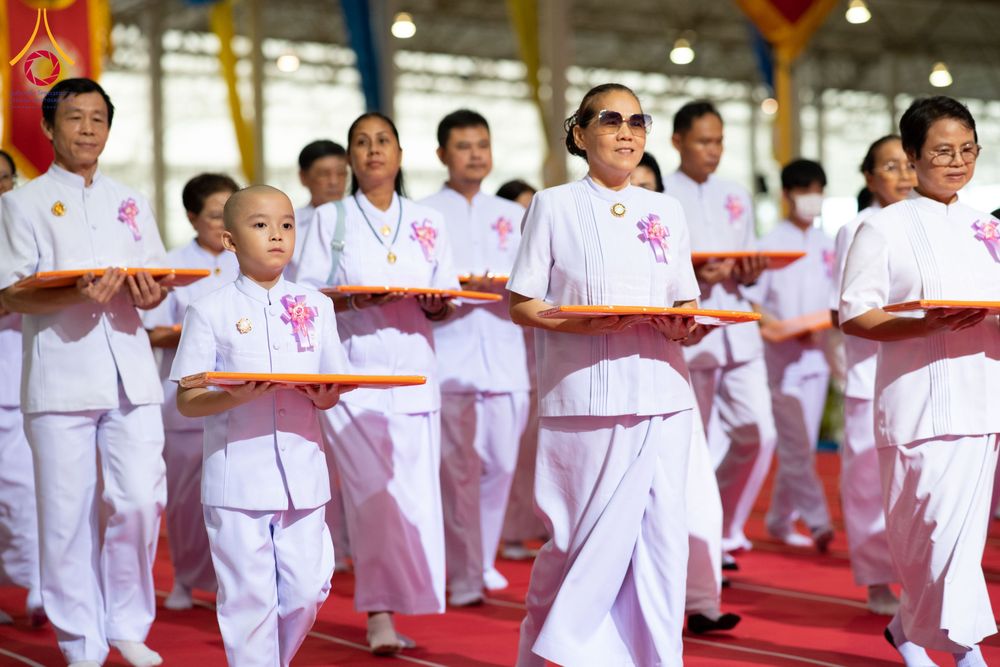กลุ่มคนสวมชุดขาวถือพานสีส้มเดินในขบวนพิธีการภายในอาคาร