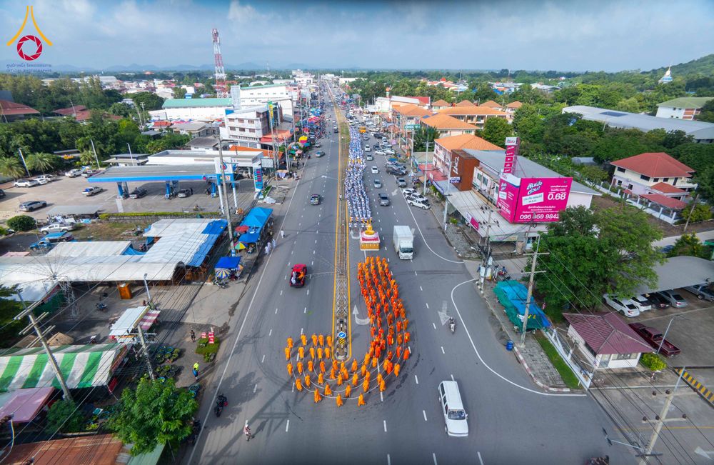 ขบวนพระภิกษุสงฆ์จำนวนมากเดินเรียงแถวบนถนนในเขตชุมชน โดยมีรถอัญเชิญพระพุทธรูปนำหน้า