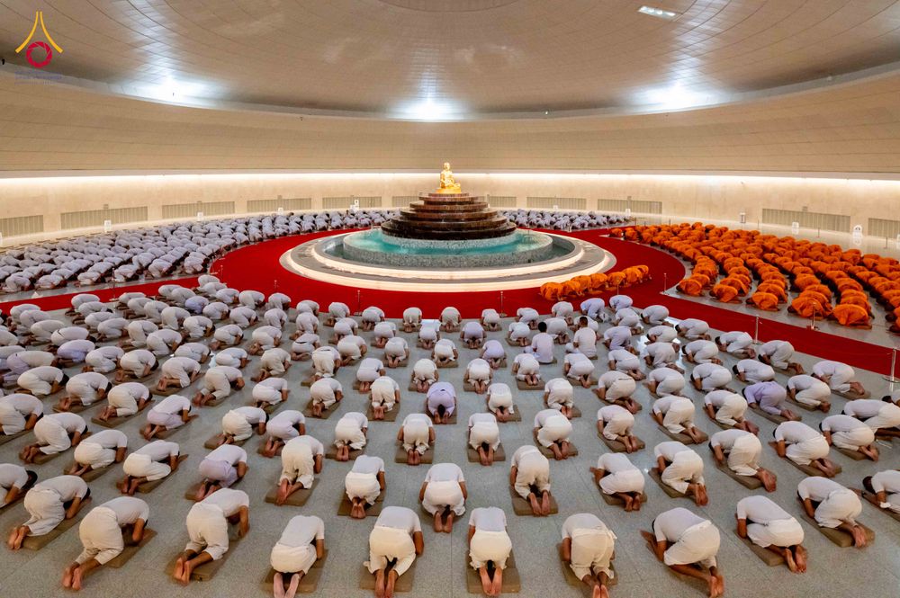 กลุ่มคนจำนวนมากในชุดสีขาวและพระภิกษุในชุดสีส้มกำลังกราบลงกับพื้นภายในอาคารทรงกลมขนาดใหญ่ที่มีพระพุทธรูปอยู่ตรงกลาง