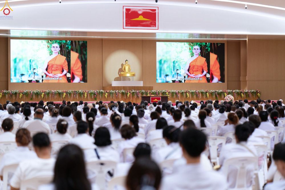 ผู้คนจำนวนมากสวมชุดสีขาวนั่งฟังการบรรยายในห้องประชุมที่มีจอภาพขนาดใหญ่และพระพุทธรูปประดิษฐานอยู่บนเวที