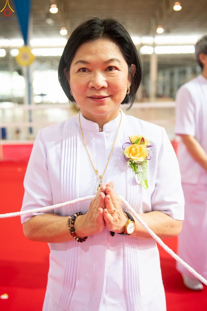 หญิงวัยกลางคนสวมชุดสีขาวพนมมือไหว้ มีดอกไม้ติดที่หน้าอกและมีเชือกสีขาวพาดผ่าน