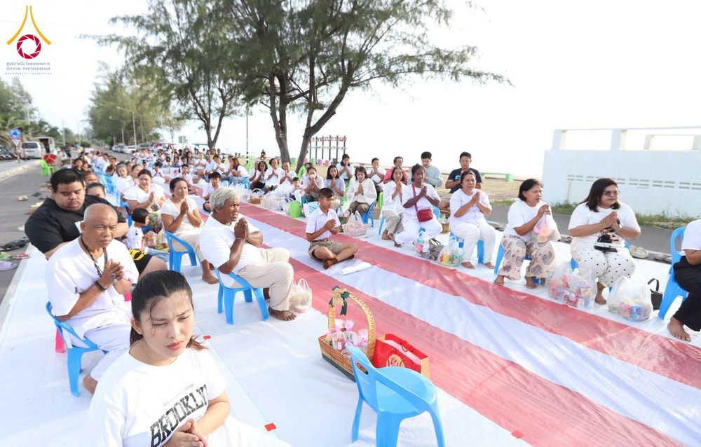 กลุ่มประชาชนสวมชุดสีขาวนั่งพนมมือเรียงแถวบนเสื่อริมชายหาดเพื่อรอตักบาตร