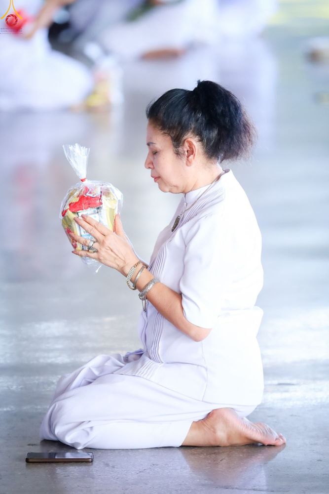 หญิงสาวสวมชุดสีขาวนั่งพับเพียบถือถุงอาหารแห้งด้วยความตั้งใจในพิธีทำบุญ