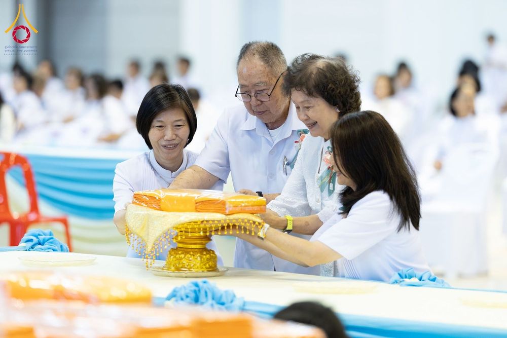 กลุ่มคนสวมชุดขาวกำลังร่วมกันประคองพานพุ่มที่มีผ้าไตรจีวรวางอยู่บนโต๊ะในพิธีทางศาสนา