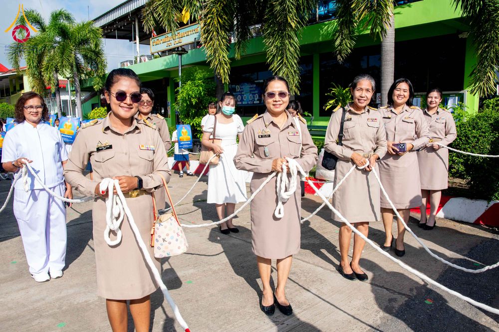 กลุ่มบุคคลในชุดข้าราชการและชุดสีขาวถือเชือกสีขาวในพิธีทางศาสนาหน้าอาคาร