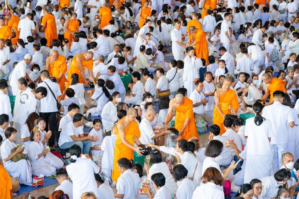 พุทธศาสนิกชนสวมชุดขาวกำลังตักบาตรข้าวสารอาหารแห้งแด่พระภิกษุสงฆ์ในอาคารขนาดใหญ่
