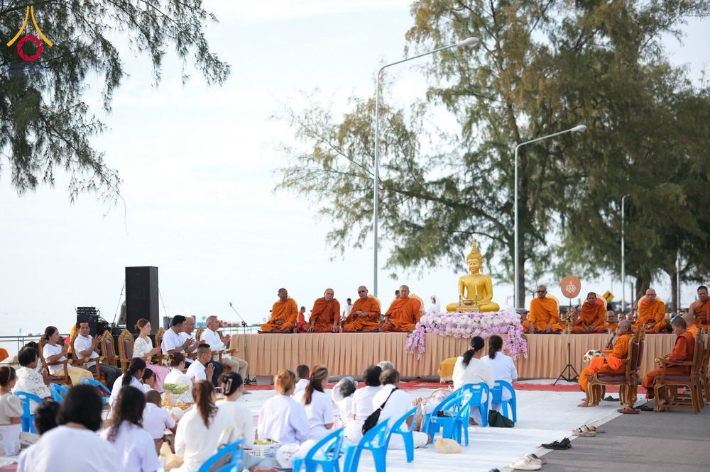 กลุ่มคนสวมชุดขาวนั่งร่วมพิธีตักบาตรพระสงฆ์บนเวทีริมชายหาด