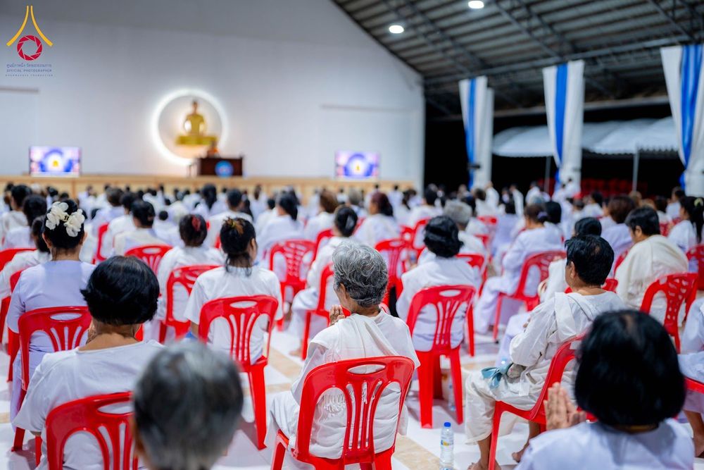 กลุ่มคนสวมชุดสีขาวนั่งเรียงแถวบนเก้าอี้สีแดงภายในหอประชุมเพื่อร่วมกิจกรรมปฏิบัติธรรม