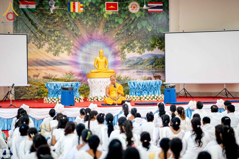 พระภิกษุสงฆ์นั่งบนเวทีแสดงธรรมต่อหน้าผู้เข้าร่วมปฏิบัติธรรมที่สวมชุดขาวจำนวนมากในห้องโถง