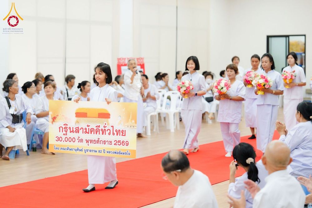 เด็กหญิงถือป้ายกฐินสามัคคีเดินนำขบวนผู้เข้าร่วมโครงการสวมชุดขาวถือพานดอกไม้ในอาคาร