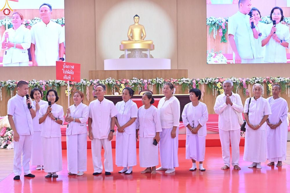 กลุ่มพุทธศาสนิกชนในชุดขาวกำลังยืนทำกิจกรรมบนเวทีภายในอาคารที่มีพระพุทธรูปเป็นฉากหลัง