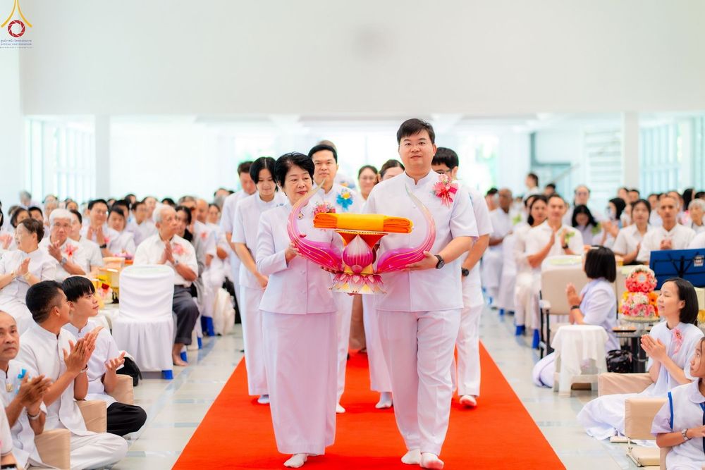 กลุ่มคนในชุดสีขาวกำลังเดินอัญเชิญพานผ้าไตรบนพรมแดงภายในอาคาร โดยมีผู้เข้าร่วมงานนั่งพนมมืออยู่โดยรอบ