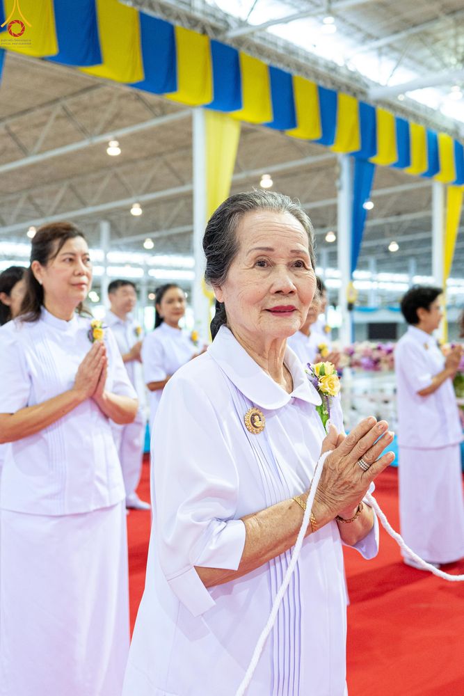 หญิงสูงวัยสวมชุดสีขาวพนมมือถือสายสิญจน์ร่วมพิธีทางศาสนาท่ามกลางพุทธศาสนิกชนในอาคาร