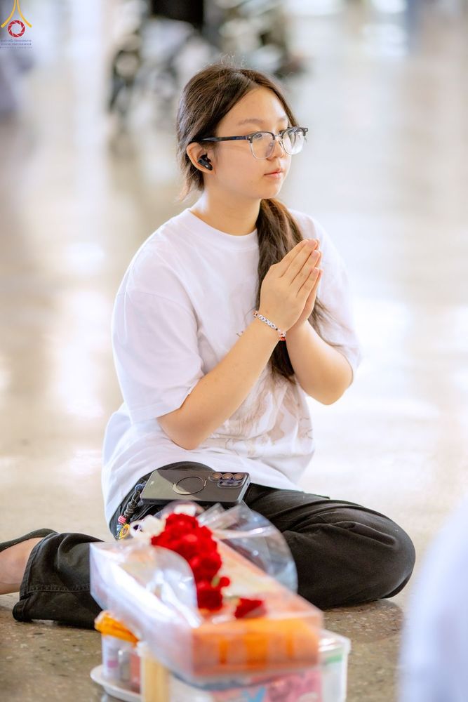 หญิงสาวสวมแว่นตาและเสื้อยืดสีขาวนั่งพนมมือไหว้ในท่าทางสงบ