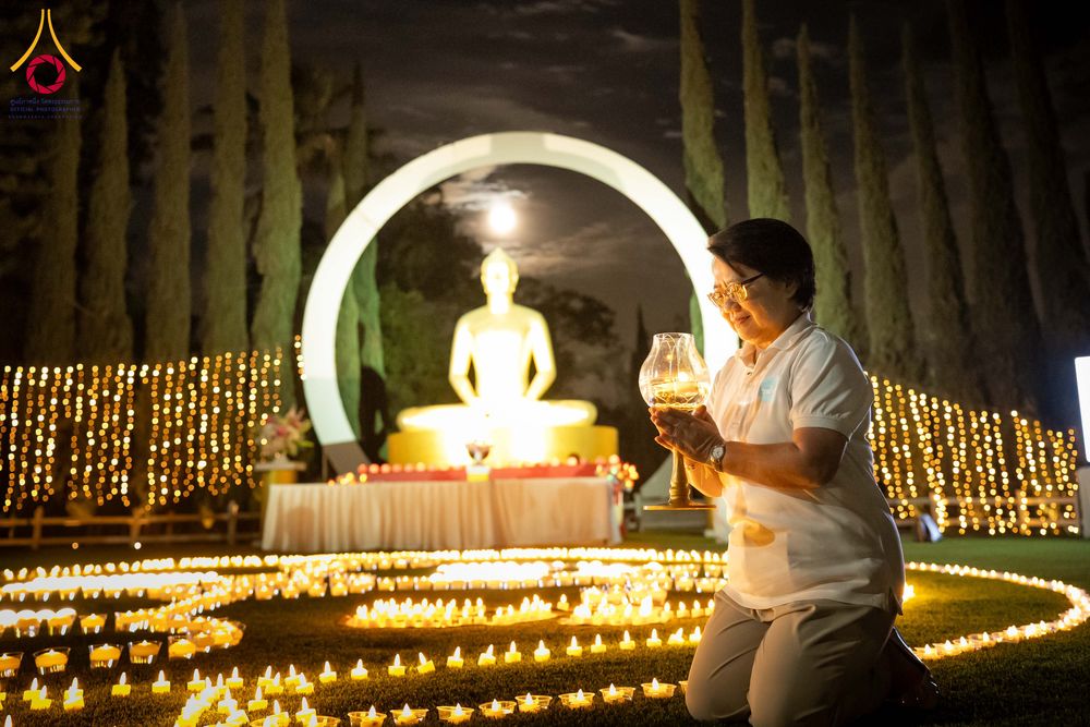 หญิงไทยถือโคมประทีปในกิจกรรมจุดประทีปท่ามกลางแสงไฟประดับและพระพุทธรูปในยามค่ำคืน
