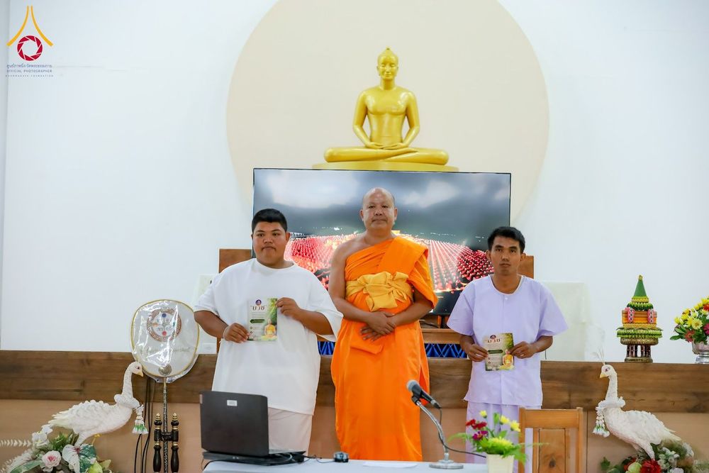 พระภิกษุและอุบาสกยืนถ่ายภาพร่วมกันในห้องอบรมที่มีพระพุทธรูปเป็นฉากหลัง