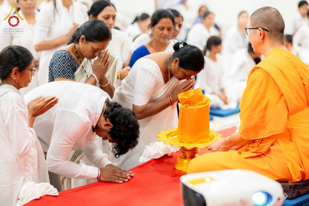 พุทธศาสนิกชนในชุดสีขาวกราบพระภิกษุสงฆ์ในจีวรสีส้มท่ามกลางบรรยากาศการทำกิจกรรมทางศาสนา
