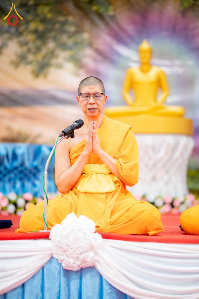 ภิกษุณีสวมแว่นตานั่งพนมมือสวดมนต์หน้าไมโครโฟนบนเวทีที่มีพระพุทธรูปเป็นฉากหลัง