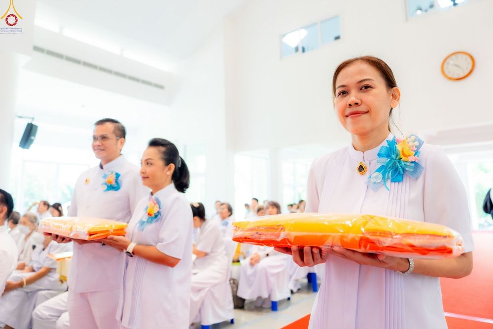 หญิงไทยในชุดสีขาวถือผ้าไตรจีวรสีเหลืองทองในงานบุญ โดยมีผู้เข้าร่วมงานคนอื่นๆ อยู่ในพื้นหลัง