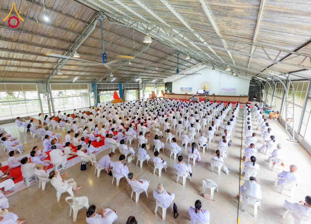 กลุ่มคนสวมชุดสีขาวนั่งเรียงแถวบนเก้าอี้พลาสติกภายในอาคารโถงกว้างเพื่อร่วมกิจกรรมอบรม