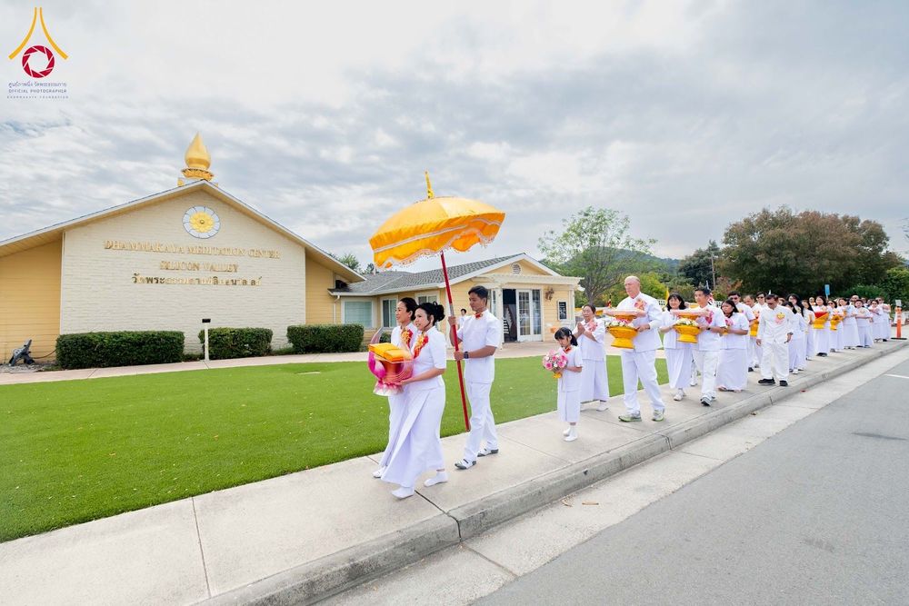 กลุ่มคนชุดขาวเดินแถวถือพานผ้าป่าและกางร่มสีเหลืองหน้าอาคารวัดพระธรรมกายซิลิคอนวัลเลย์
