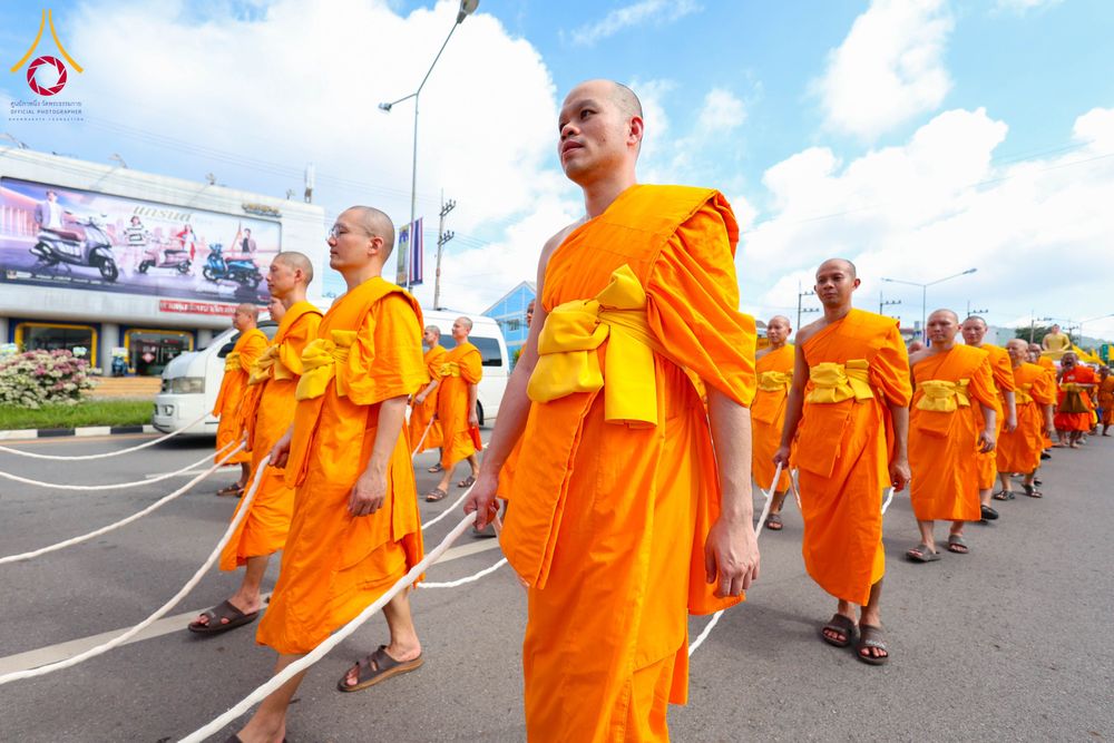 คณะสงฆ์หลายรูปสวมจีวรสีส้มเดินเรียงแถวบนถนนและถือสายสิญจน์ในมือ