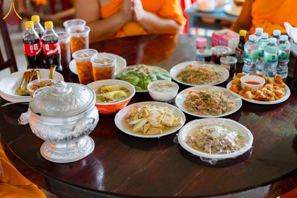 อาหารไทยหลายเมนูวางบนโต๊ะไม้กลมพร้อมเครื่องดื่มและพระสงฆ์นั่งล้อมรอบ