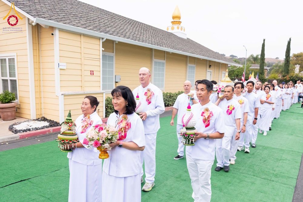 กลุ่มคนสวมชุดขาวเดินแถวในพิธีทางศาสนา โดยมีผู้ถือพานดอกไม้และพานพุ่มนำขบวนบนทางเดินสีเขียว