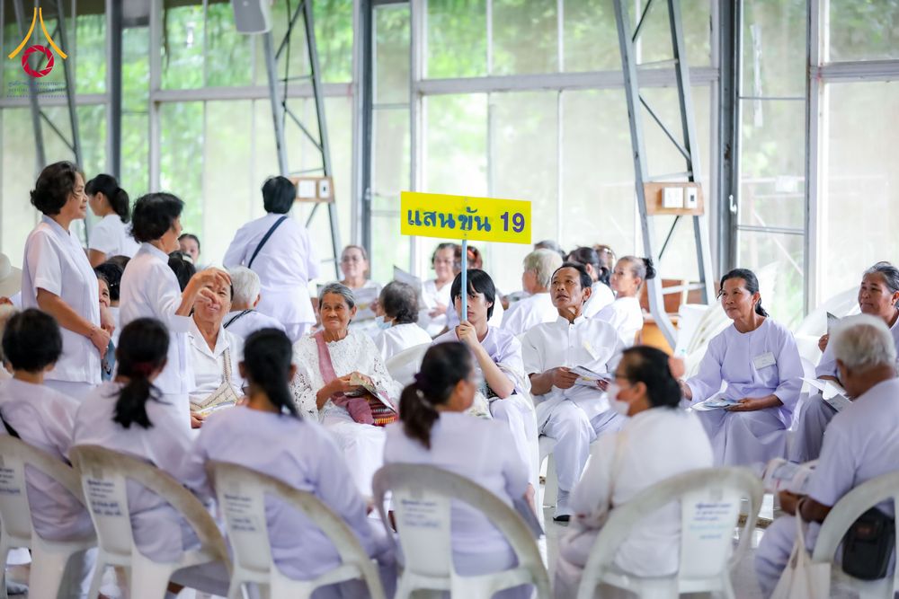 กลุ่มคนสวมชุดสีขาวนั่งทำกิจกรรมร่วมกันภายในอาคาร มีป้ายสีเหลืองเขียนว่า แสนขัน 19