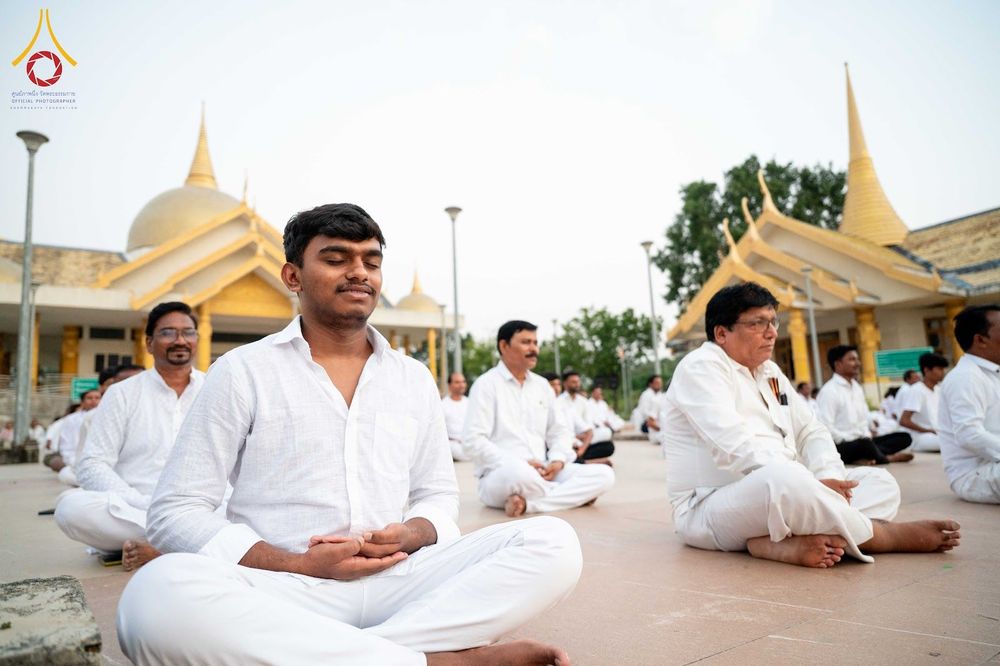 กลุ่มคนสวมชุดสีขาวนั่งสมาธิบนลานกว้างหน้าอาคารทรงไทยในประเทศอินเดีย