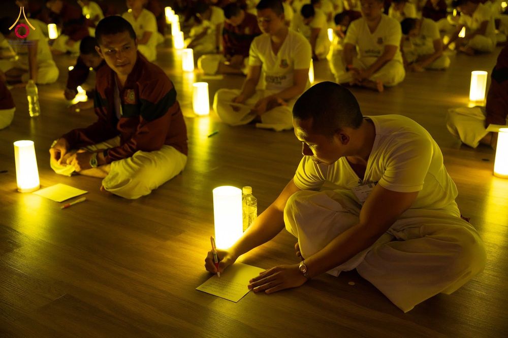 กลุ่มคนสวมชุดสีขาวนั่งปฏิบัติธรรมบนพื้นไม้ท่ามกลางแสงไฟจากโคมไฟทรงกระบอกในห้องโถง