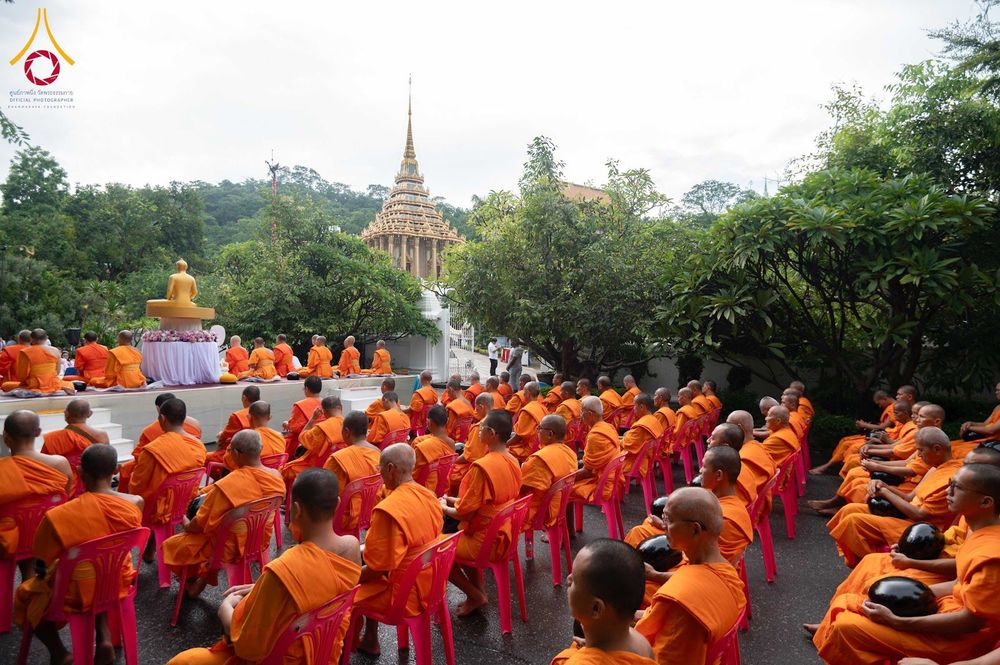 สามเณรจำนวนมากนั่งบนเก้าอี้สีชมพูในพิธีตักบาตร ณ วัดที่มีพระพุทธรูปและต้นไม้