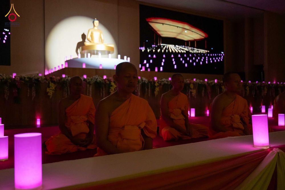 พระสงฆ์นั่งสมาธิเรียงแถวหน้าพระพุทธรูปในห้องโถงที่มีแสงไฟสีชมพูและเทียนประดับ