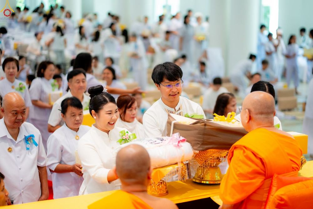กลุ่มคนสวมชุดขาวกำลังถวายสิ่งของแด่พระสงฆ์ในงานบุญที่มีผู้คนมาร่วมงานจำนวนมาก