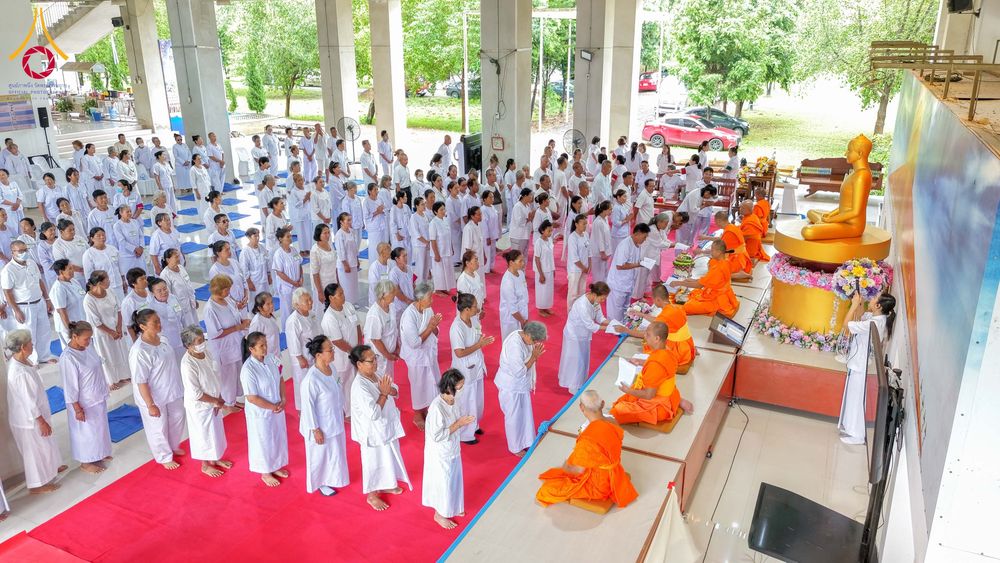 กลุ่มคนในชุดขาวจำนวนมากกำลังร่วมกิจกรรมทางศาสนาโดยมีพระภิกษุสงฆ์นั่งบนเวทีด้านหน้าพระพุทธรูป