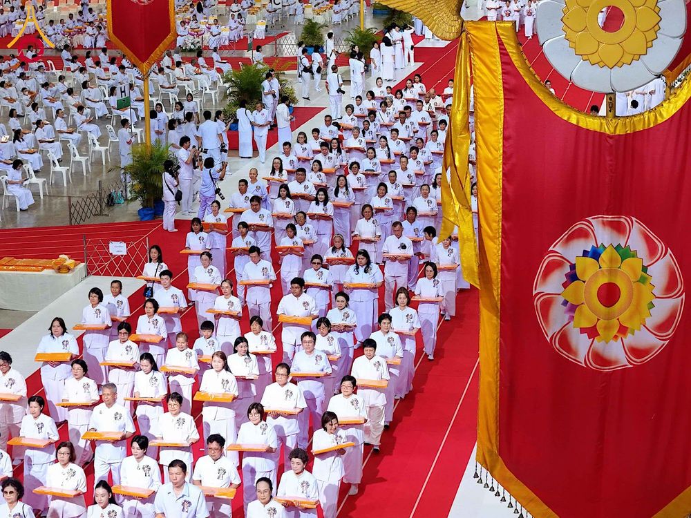 กลุ่มพุทธศาสนิกชนสวมชุดสีขาวถือพานผ้าไตรจีวรยืนเรียงแถวในพิธีทางศาสนาภายในอาคารขนาดใหญ่