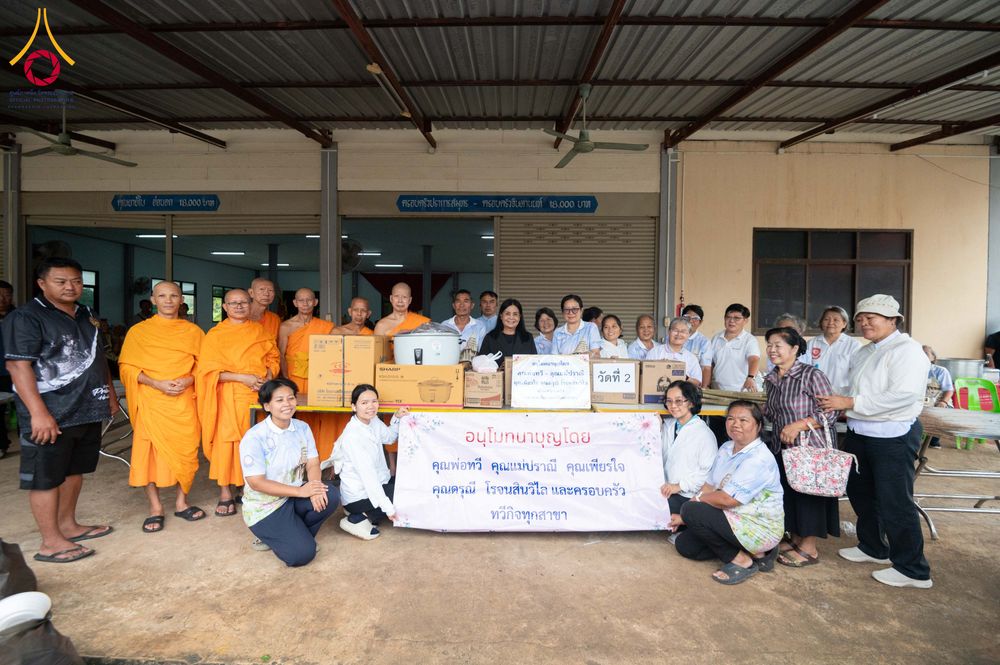 พระภิกษุและกลุ่มคนยืนถ่ายภาพร่วมกันหน้ากล่องสิ่งของบริจาคและป้ายผ้าที่มีข้อความระบุชื่อผู้สนับสนุนการบริจาค