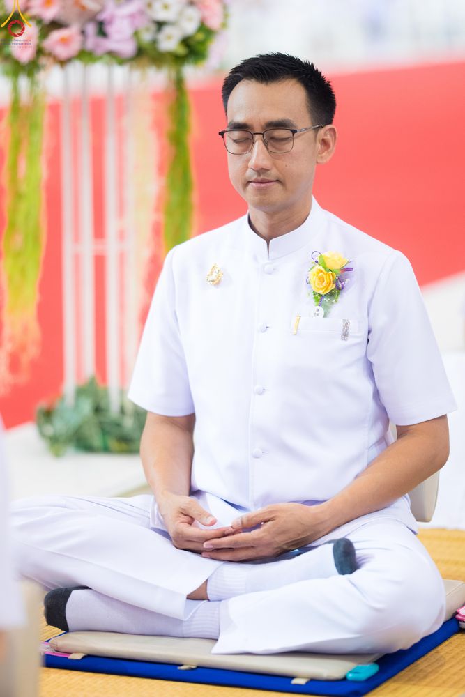 ชายสวมชุดขาวนั่งสมาธิหลับตาอย่างสงบ มีดอกไม้ประดับที่หน้าอก