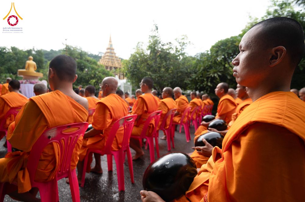 สามเณรหลายรูปในจีวรสีส้มถือบาตรสีดำนั่งเรียงแถวบนเก้าอี้พลาสติกสีชมพูในบริเวณวัด