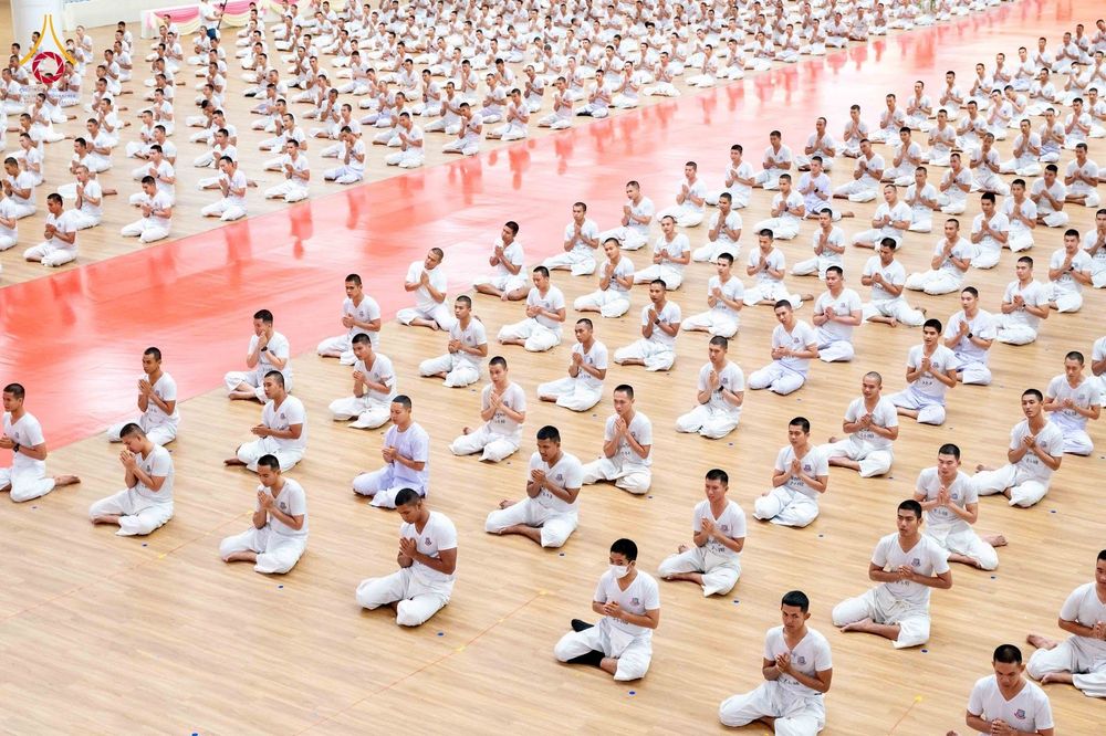 นักเรียนนายสิบตำรวจจำนวนมากสวมชุดสีขาวนั่งพนมมือบนพื้นไม้ในหอประชุม