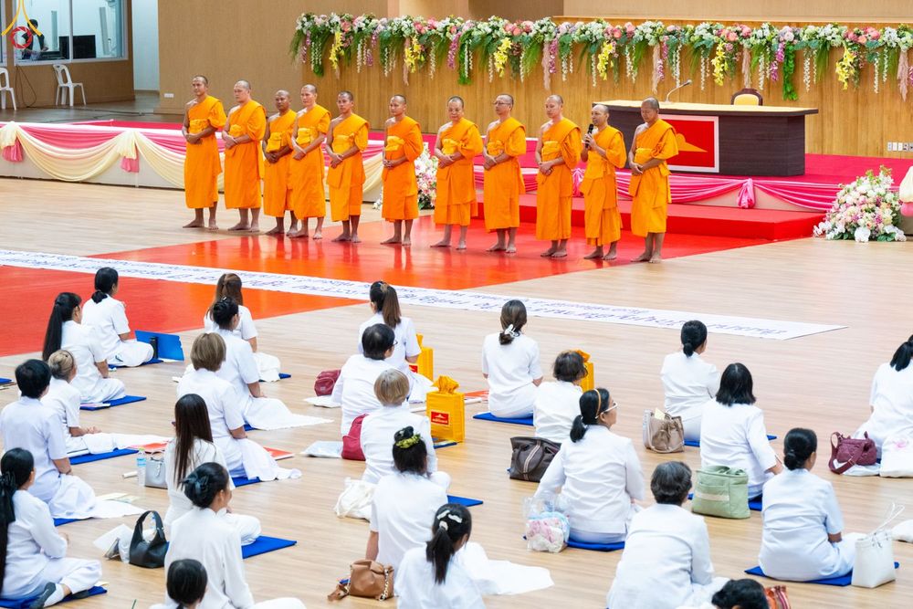 พระภิกษุสงฆ์ยืนบนเวทีและสาธุชนชุดขาวนั่งปฏิบัติธรรมในหอประชุม