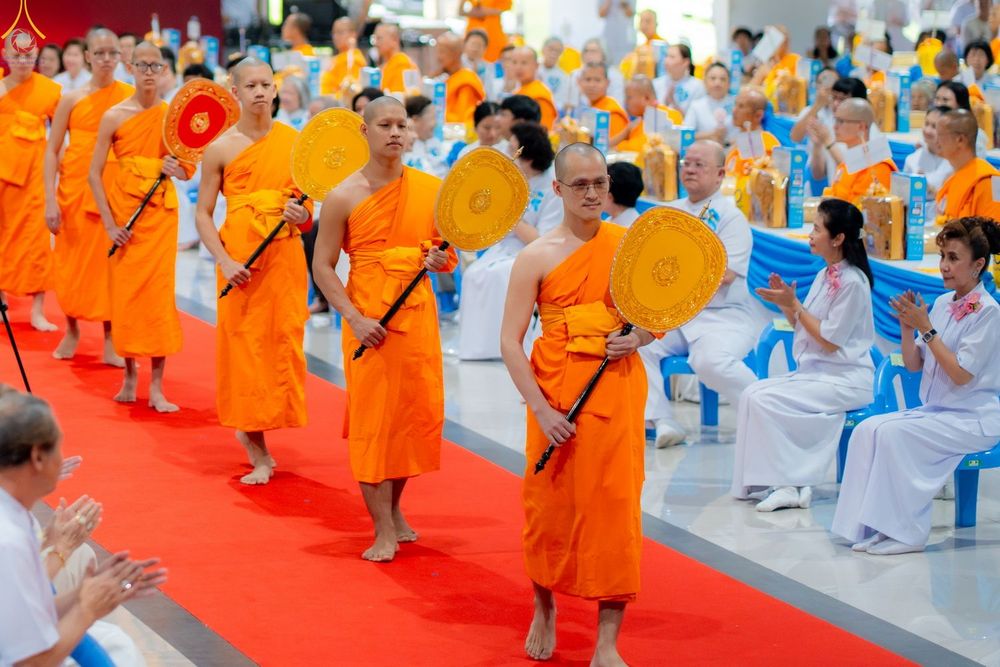 พระสงฆ์หลายรูปในจีวรสีส้มถือพัดยศเดินเรียงแถวบนพรมสีแดงท่ามกลางพุทธศาสนิกชนที่นั่งพนมมือ