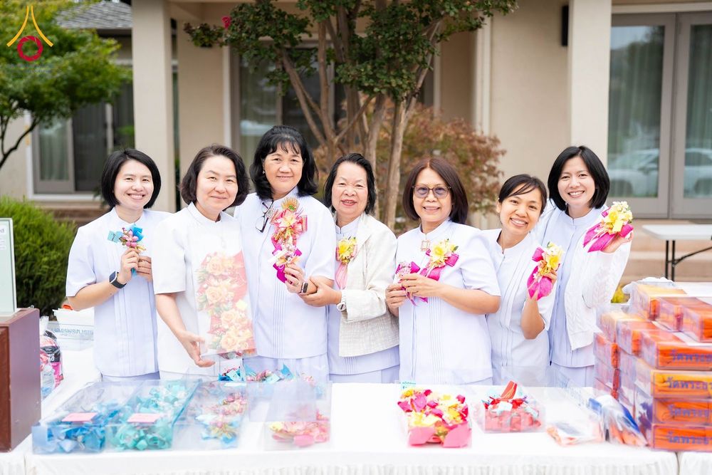 กลุ่มผู้หญิง 7 คนสวมชุดสีขาวถือดอกไม้ประดิษฐ์ยืนยิ้มหน้าโต๊ะที่มีของตกแต่งและกล่องของขวัญ