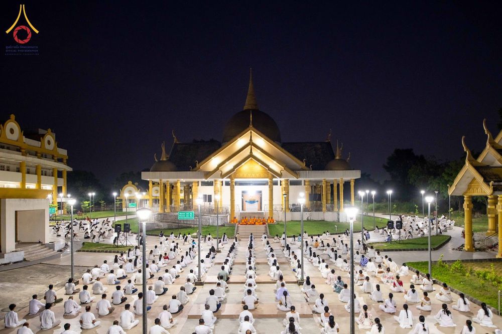 กลุ่มคนสวมชุดสีขาวนั่งสมาธิบนลานกว้างหน้าอาคารทรงไทยในยามค่ำคืน